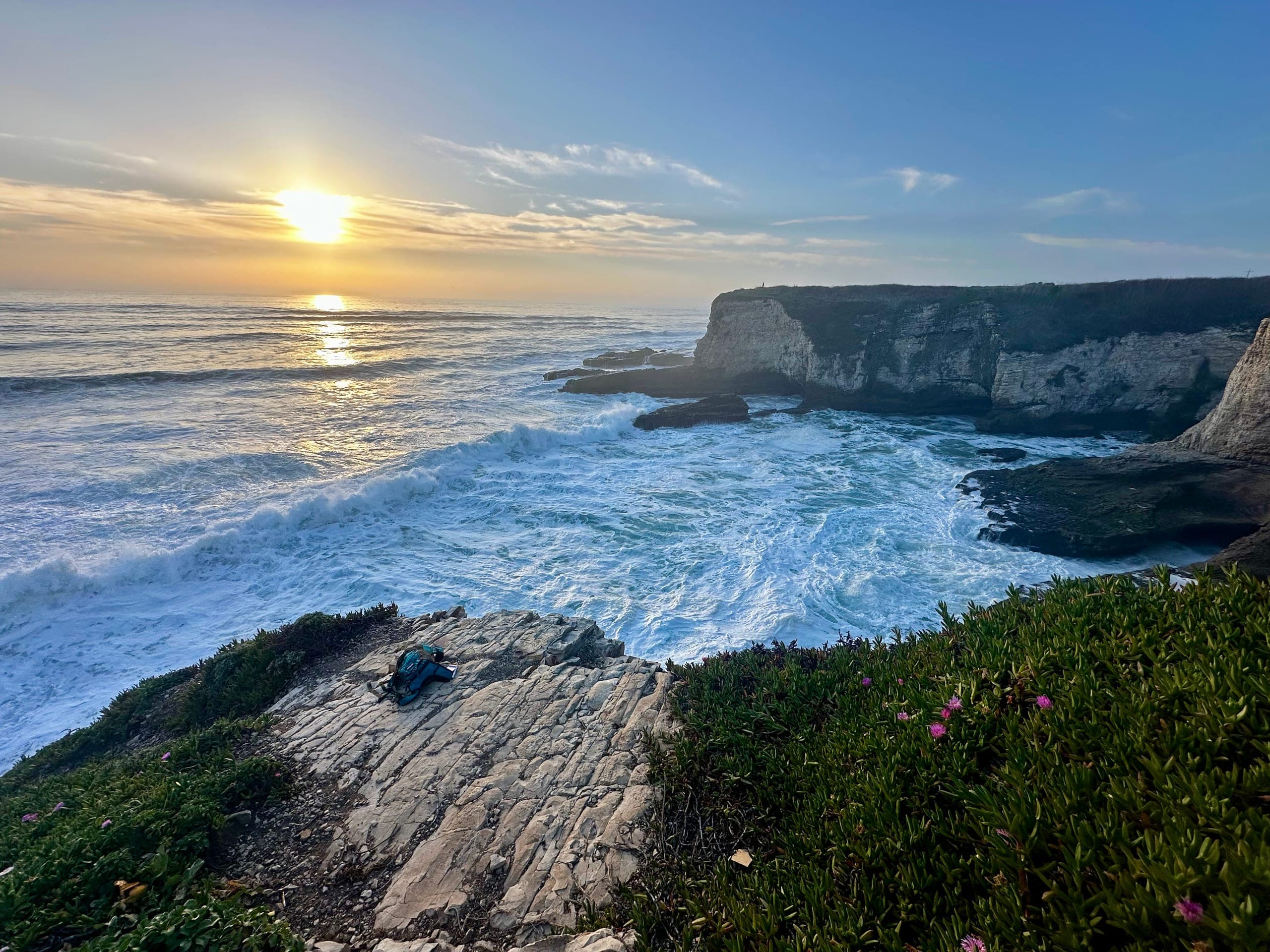 Sunset over a rocky Pacific coastline, white surf breaking beneath the cliffs